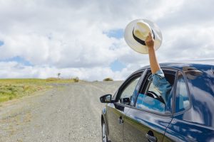 A passenger in the backseat with their arm out the window holding a hat. Driving on a dirt road.